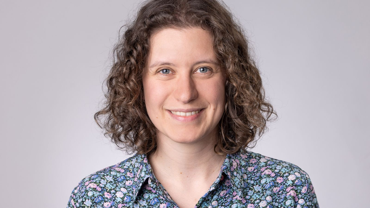 Lächelnde Person mit lockigem Haar und floralem Hemd vor neutral grauem Hintergrund.