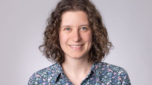 Lächelnde Person mit lockigem Haar und floralem Hemd vor neutral grauem Hintergrund.