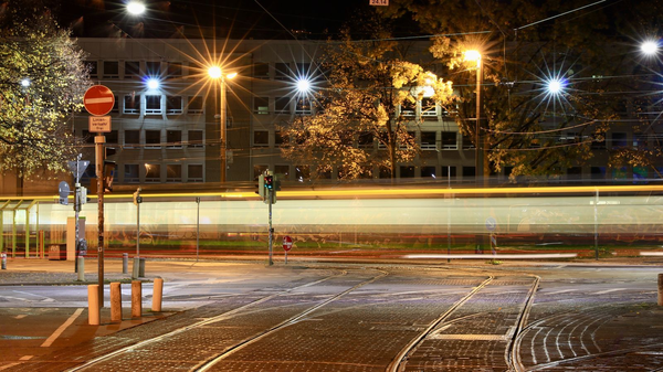 Nächtliche Straßenaufnahme mit vorbeifahrender Straßenbahn, Schienen im Vordergrund und sternförmigen Lichtern.