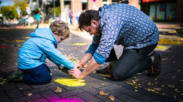 Erwachsener und Kind sprühen gemeinsam gelbe Farbe auf eine Straße im Freien.