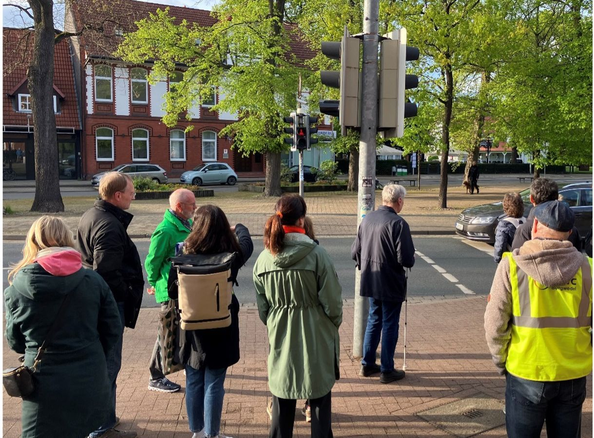 Gruppe von Menschen steht an einer Ampelkreuzung und wartet, im Hintergrund rote Backsteingebäude und grüne Bäume.