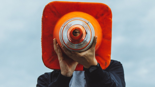 Person hält einen orangefarbenen Verkehrskegel vor das Gesicht, durch dessen Öffnung ein Auge sichtbar ist.