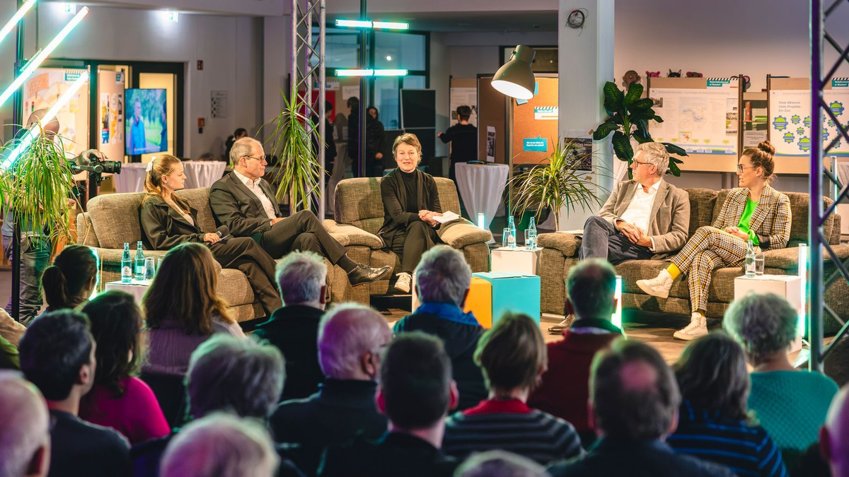 Podiumsdiskussion mit fünf Personen auf Sofas vor Publikum in modern beleuchtetem Raum.