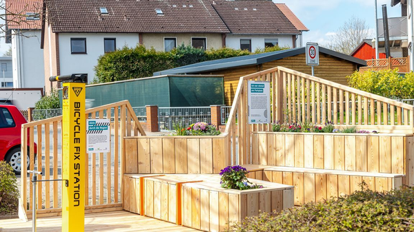 Holzinstallation mit Sitzflächen, Blumenbeeten und gelber Fahrradreparaturstation, Schild „Ich war einmal ein Parkplatz“.