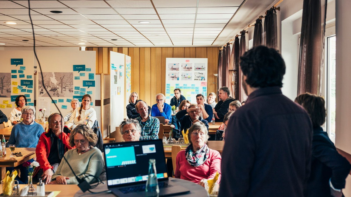 Teilnehmende eines Workshops hören einer Präsentation in einem Raum mit Postern und Haftnotizen zu.