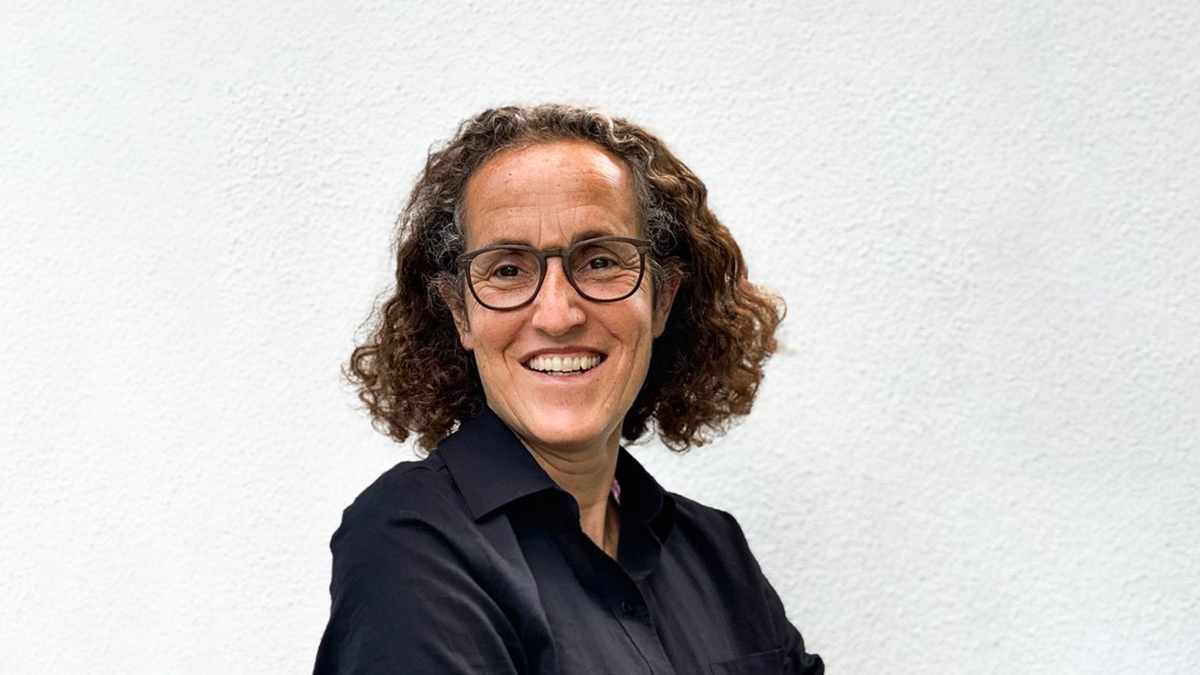 Porträt einer lachenden Person mit Brille und dunklem Hemd vor einer weißen Wand.