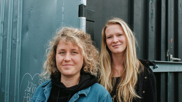 Zwei Frauen stehen nebeneinander vor einer blaugrauen Metallwand und blicken in die Kamera.