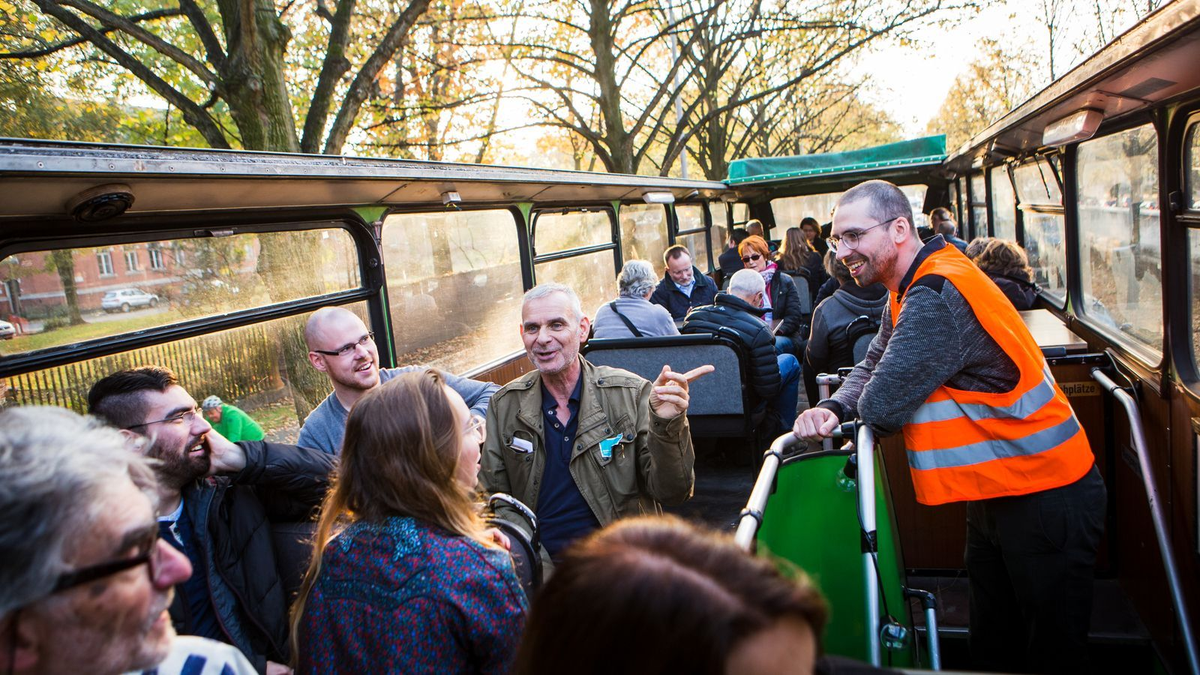 Menschen sitzen in einem offenen Bus und unterhalten sich, ein Mann mit Warnweste steht im Gang, herbstliche Sonne scheint durch die Bäume.