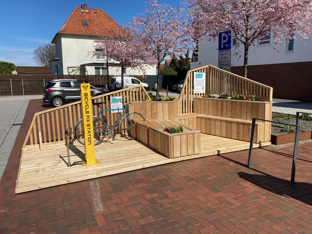 Ein hölzernes Parklet mit Sitzbänken, Blumenbeeten und einer gelben Fahrradreparaturstation steht am Straßenrand; daneben ein Fahrrad, im Hintergrund blühende Bäume und Wohnhäuser.