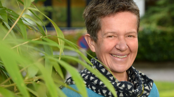 Lächelnde Person mit kurzem Haar und Schal steht im Freien neben grünen Pflanzen.