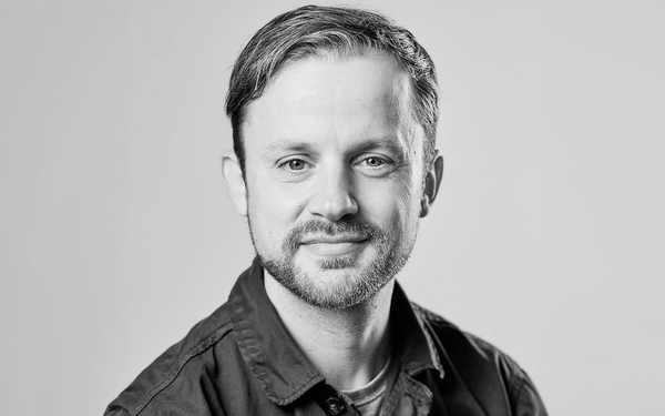 Schwarz-weiß-Porträt eines Mannes mit kurzem Haar, Bart und offenem Hemd, freundlich in die Kamera blickend.