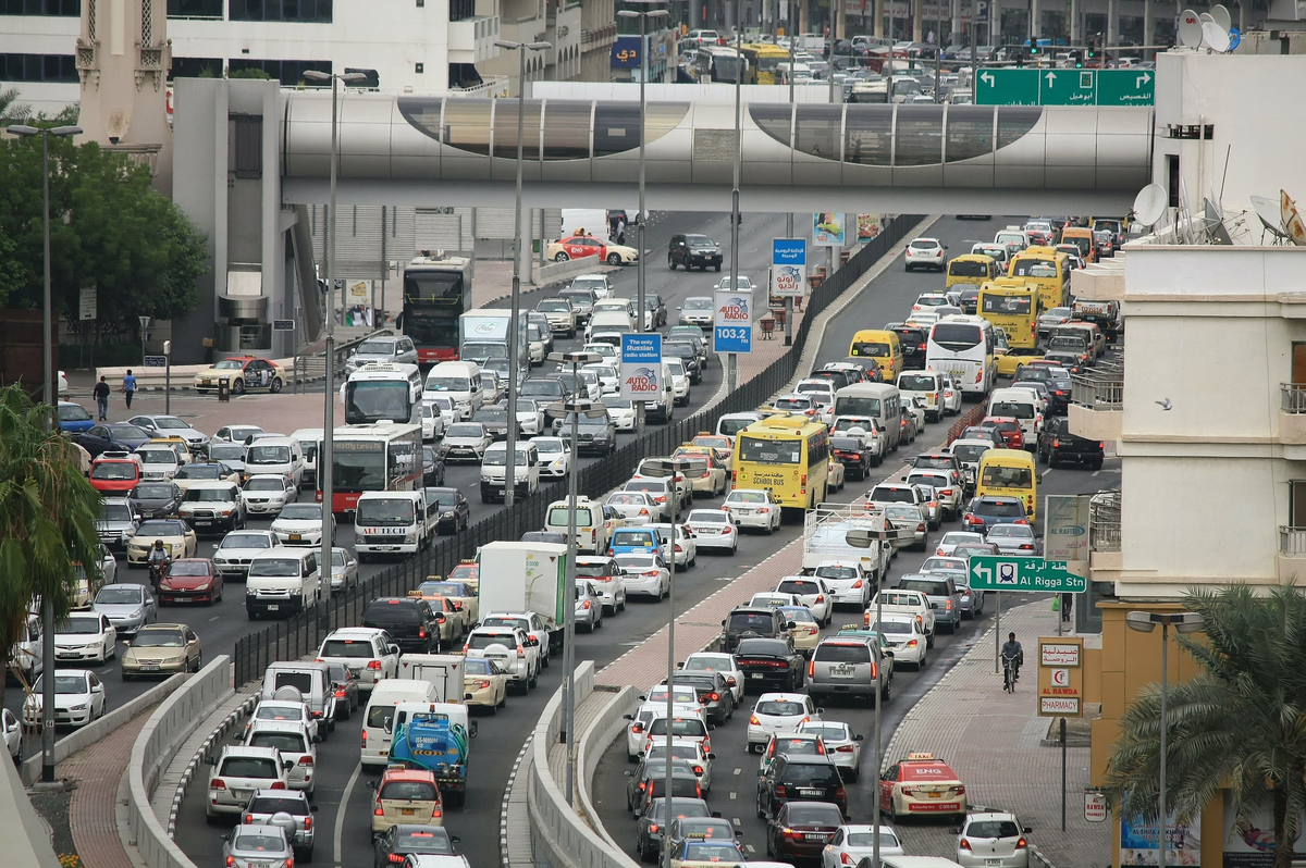 Verkehrsstau auf einer mehrspurigen Stadtstraße mit Autos, Bussen und Hochhäusern im Hintergrund.