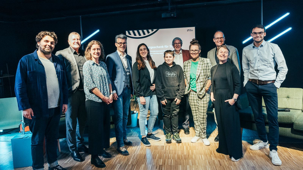 Gruppenfoto von zwölf Personen auf einer Bühne vor einer Leinwand bei einer Veranstaltung, freundlich in die Kamera blickend.