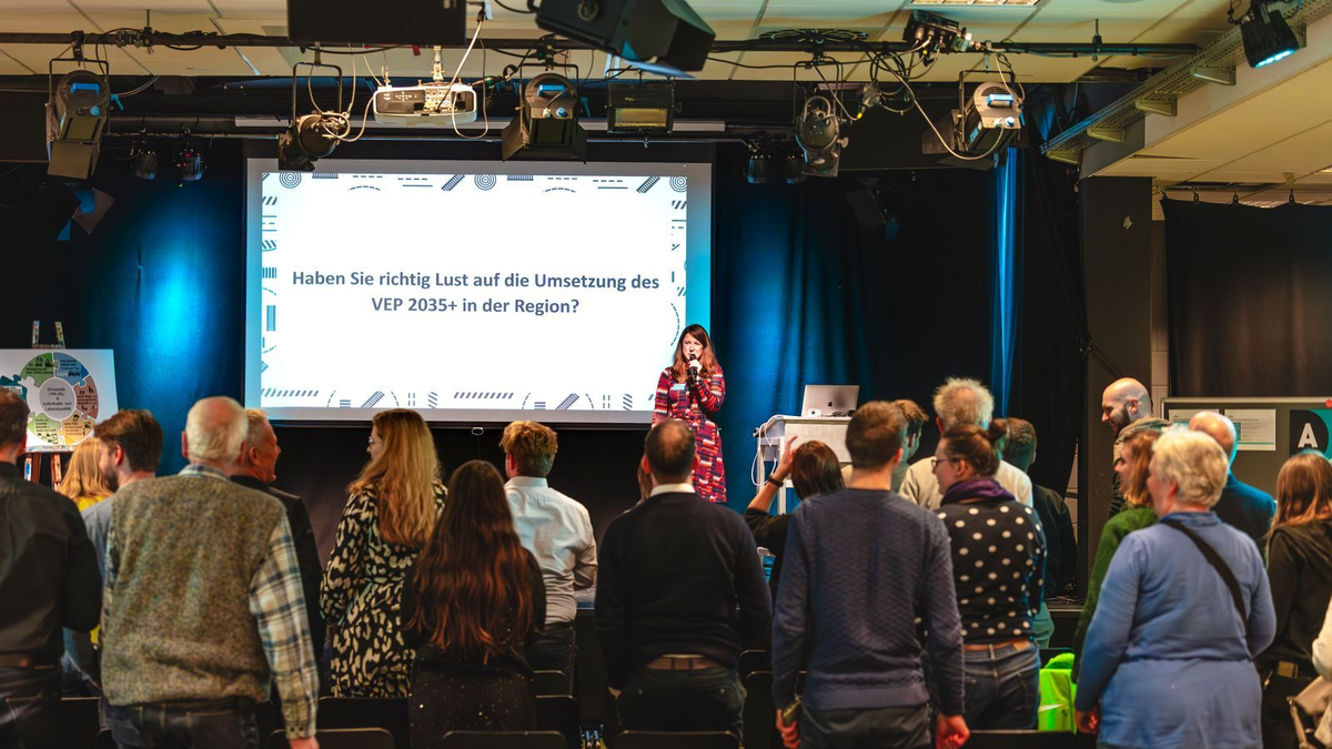 Frau spricht auf Bühne vor Publikum, hinter ihr steht auf Leinwand „Haben Sie richtig Lust auf die Umsetzung des VEP 2035+ in der Region?“.