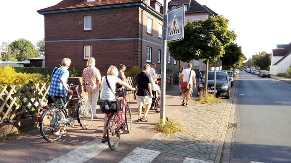 Gruppe von Menschen mit Fahrrädern und Rollstuhl auf einem Gehweg entlang einer Wohnstraße bei sonnigem Wetter.