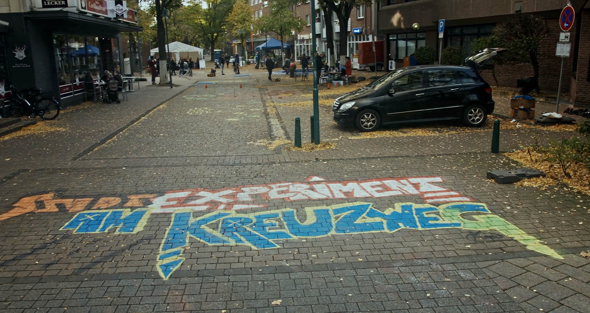 Bunte Straßenbemalung mit der Aufschrift „Stadtexperimente am Kreuzweg“ in einer herbstlichen Innenstadtstraße.